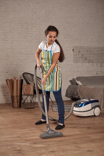 Jeune femme utilisant un aspirateur dans le sol du salon, effectuant des tâches de nettoyage et des tâches ménagères, intérieur méticuleux.