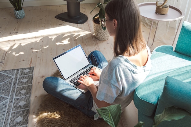 Jeune femme utilisant un écran blanc pour ordinateur portable pour travailler, assise par terre à la maison. Horaires flexibles et télétravail.