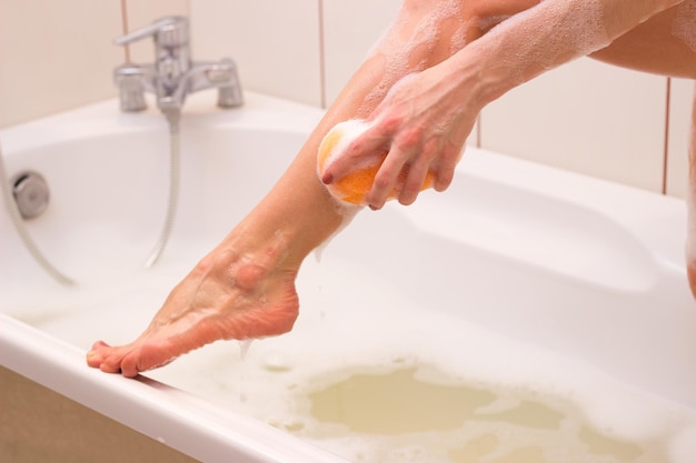 Jeune femme utilisant une éponge orange pour laver sa jambe dans la baignoire blanche pleine de mousse dans sa salle de bain