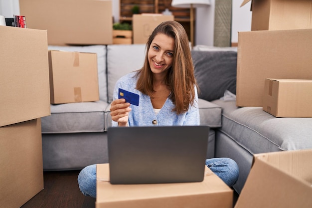 Jeune femme utilisant un ordinateur portable et une carte de crédit assise sur le sol dans une nouvelle maison