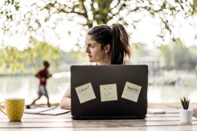 Jeune femme utilisant un ordinateur portable dans le parc public Freelance du millénaire travaillant à distance et prenant des notes dans un parc de la ville Technologie et concept de travail à distance Utilisation d'appareils technologiques à l'extérieur