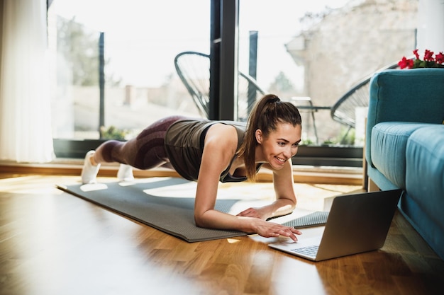 Jeune femme utilisant un ordinateur portable tout en faisant des exercices de planche à la maison le matin.