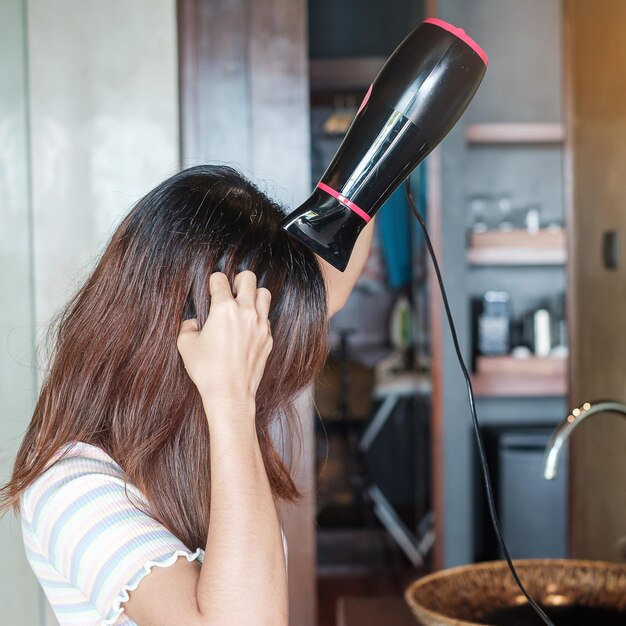 Jeune femme utilisant un sèche-cheveux près d'un miroir à la maison ou à l'hôtel Coiffures et concepts de style de vie