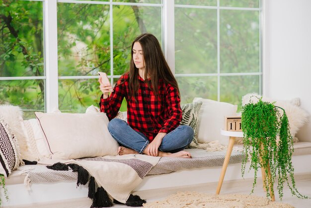 Jeune femme utilisant un smartphone près d'une grande fenêtre à la maison pendant la quarantaine