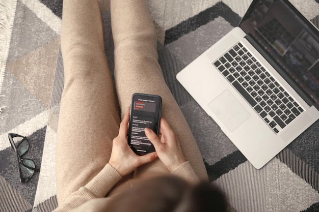 Jeune femme utilisant son téléphone portable tout en travaillant avec un ordinateur portable assis sur le sol à la maison