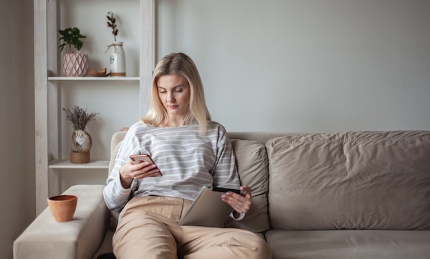 Jeune femme utilisant une tablette et un téléphone portable, assise sur un canapé à ho