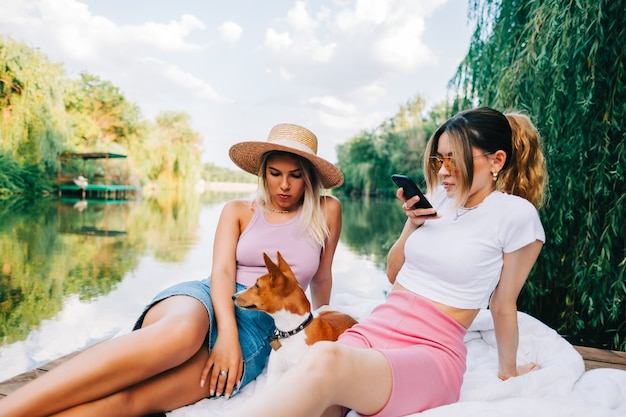 Jeune femme utilisant un téléphone portable tout en se reposant en plein air dans la nature près du lac avec des amis et un chien.