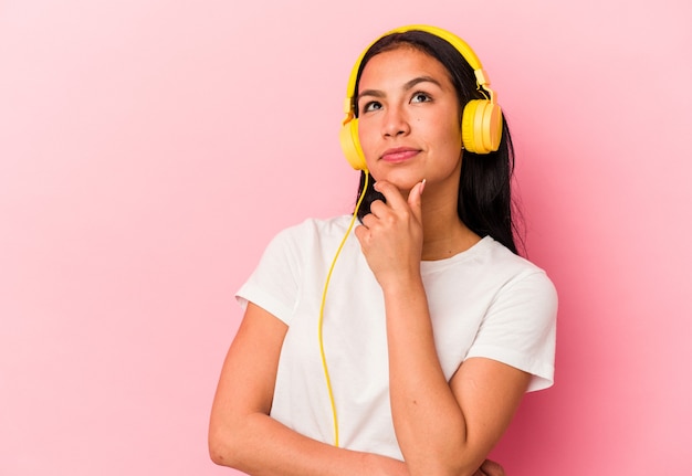 Jeune femme vénézuélienne écoutant de la musique isolée sur un mur rose regardant de côté avec une expression douteuse et sceptique.