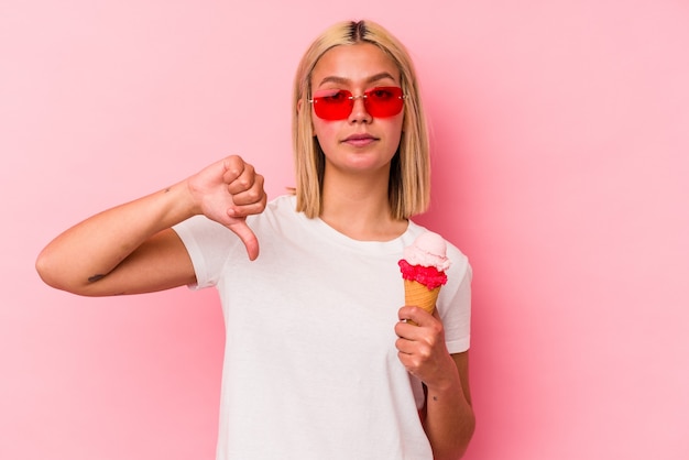 Jeune femme vénézuélienne mangeant une glace isolée sur un mur rose montrant un geste d'aversion, les pouces vers le bas. Concept de désaccord.