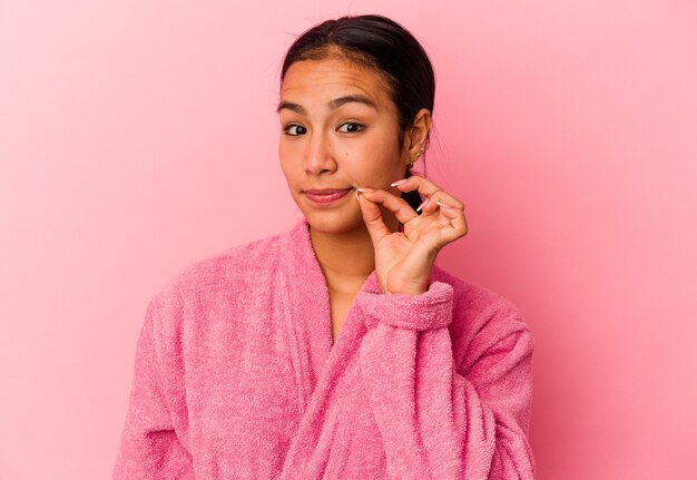 Jeune femme vénézuélienne portant un peignoir isolé sur fond rose avec les doigts sur les lèvres gardant un secret.