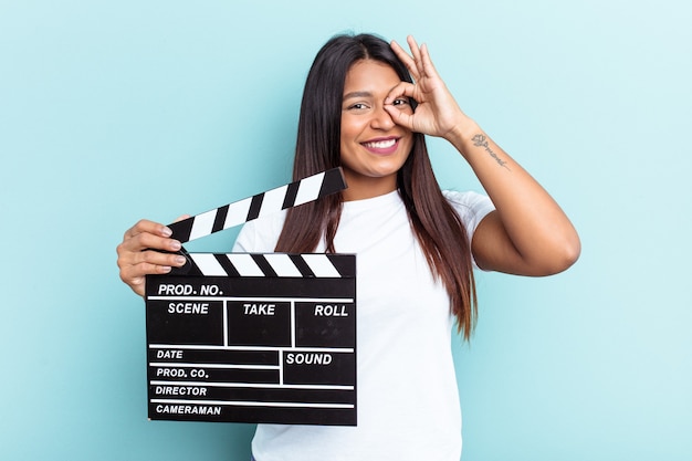 Jeune femme vénézuélienne tenant un clap isolé sur fond bleu excité en gardant le geste ok sur les yeux.