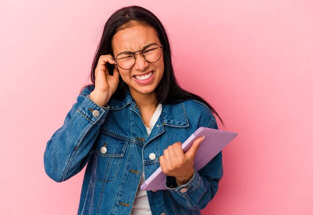 Jeune femme vénézuélienne tenant une tablette isolée sur fond rose couvrant les oreilles avec les mains.