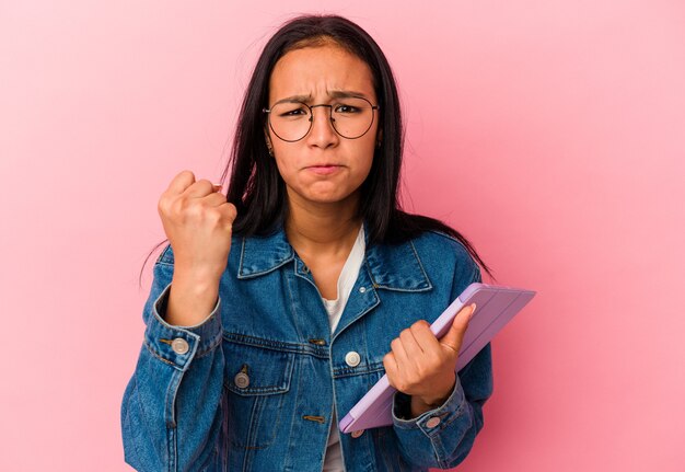 Jeune femme vénézuélienne tenant une tablette isolée sur fond rose montrant le poing à la caméra, expression faciale agressive.