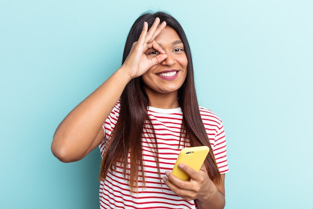 Jeune Femme Vénézuélienne Tenant Un Téléphone Portable Isolé Sur Fond Bleu Excité En Gardant Un Geste Ok Sur Les Yeux.