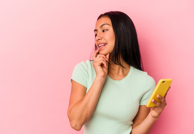 Jeune femme vénézuélienne tenant un téléphone portable isolé sur fond rose détendue en pensant à quelque chose en regardant un espace de copie.