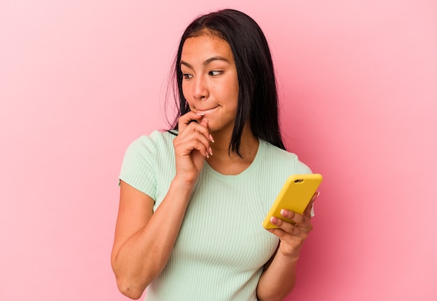 Une jeune femme vénézuélienne tenant un téléphone portable isolé sur un mur rose confuse, se sent dubitative et incertaine.