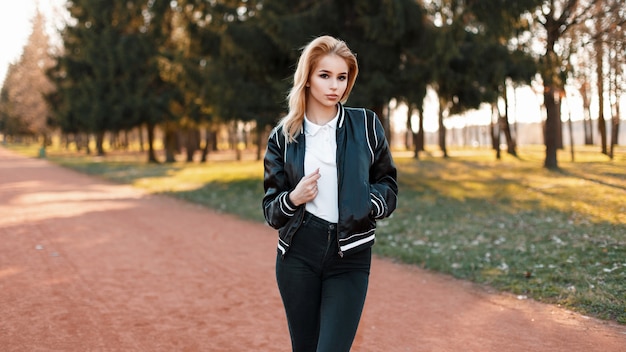 Jeune femme en veste à la mode et polo blanc et jeans noirs dans le parc par une journée ensoleillée