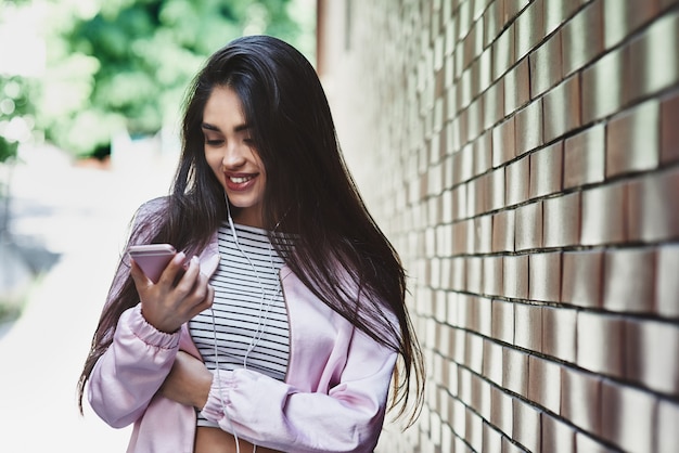 Jeune femme en veste rose debout près du mur écoutant de la musique dans