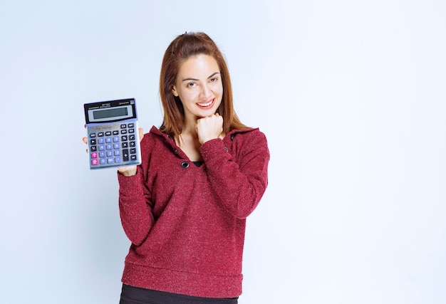 Jeune femme en veste rouge calculant quelque chose sur une calculatrice bleue et démontrant le résultat final