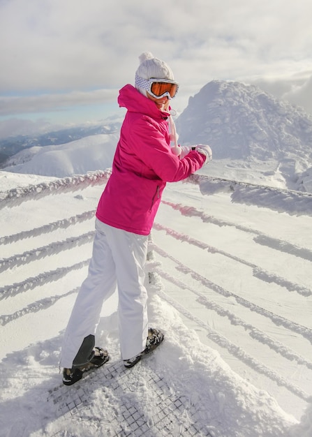 Jeune femme en veste de ski rose, bottes de pantalon, chapeau et gants, s'appuyant sur un rail couvert de neige, regardant en arrière, souriant.