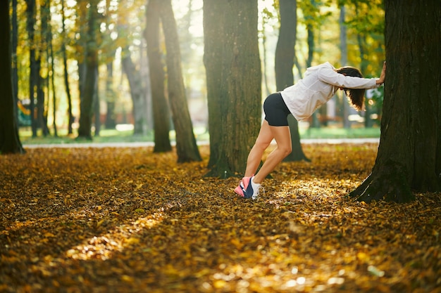 Jeune femme en veste de sport blanche et short qui s'étend du corps près de l'arbre au parc. Brune sportive faisant des exercices du matin à l'extérieur. Concept de mode de vie sain et actif.