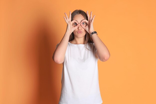 Jeune femme en vêtements de base souriante regardant la caméra à travers des trous faits avec des doigts isolés sur fond jaune.