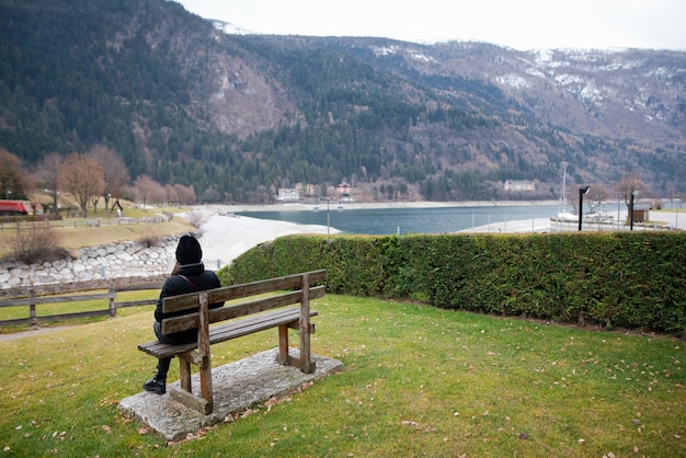 La jeune femme en vêtements chauds assis seule sur un banc en bois et regardant le lac par temps froid.