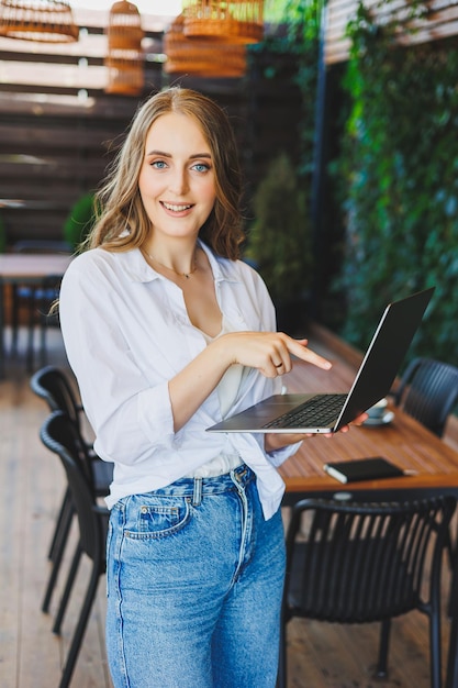 Jeune femme en vêtements décontractés buvant du café et parlant au téléphone travaillant sur une terrasse verte à un ordinateur portable regardant l'écran et travaillant sur un projet à distance du travail dans un café