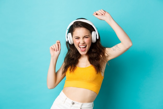 Jeune femme en vêtements d'été écoutant de la musique, portant des écouteurs et chantant la chanson préférée, dansant dans des écouteurs, debout sur fond bleu.