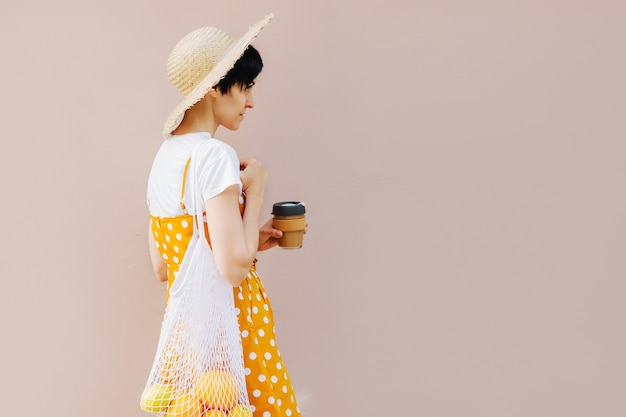 Jeune femme en vêtements d'été jaune avec un sac de fruits Eco et une tasse à café réutilisable.