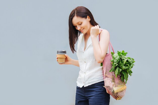 Jeune femme en vêtements d'été légers avec un sac écologique de légumes, de légumes verts et une tasse à café réutilisable. Mode de vie durable. Concept écologique.