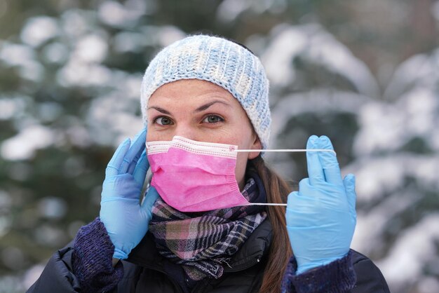 Jeune femme en vêtements d'hiver chauds, portant des gants de protection, mettant ou enlevant un masque buccal jetable à usage unique pour le virus du visage rose, gros plan, fond d'arbres couverts de neige floue