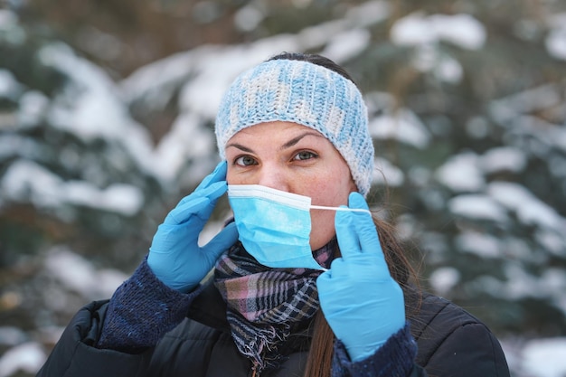 Jeune femme en vêtements d'hiver chauds, portant des gants de protection, mettant ou retirant un masque buccal jetable à usage unique pour le virus du visage bleu, gros plan, fond d'arbres couverts de neige floue