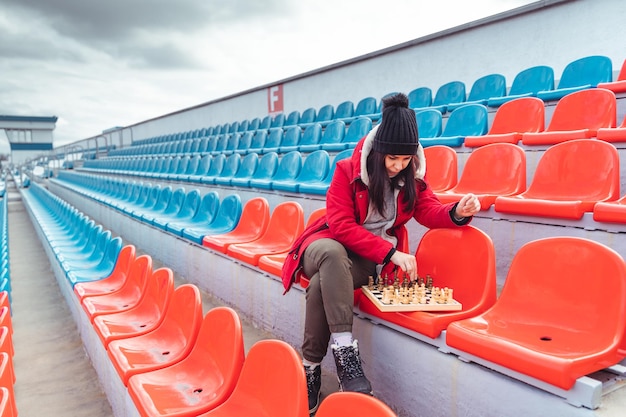 Jeune femme en vêtements d'hiver joue aux échecs assis sur les gradins du stade seul