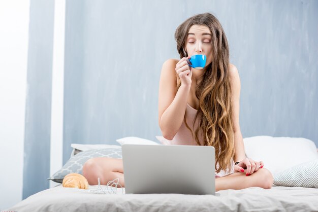 Jeune femme en vêtements de nuit buvant du café assis avec un ordinateur portable dans le lit