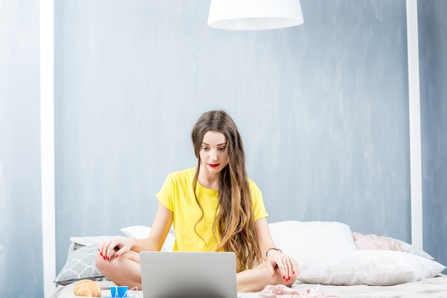 Jeune femme en vêtements de nuit travaillant avec téléphone et ordinateur portable sur le lit