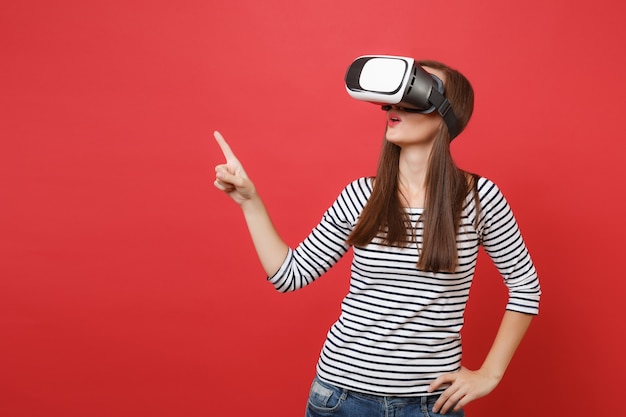 Jeune femme en vêtements à rayures décontractées lunettes de réalité virtuelle pointant l'index regardant de côté isolé sur fond de mur rouge vif. Concept de mode de vie des émotions sincères des gens. Maquette de l'espace de copie.