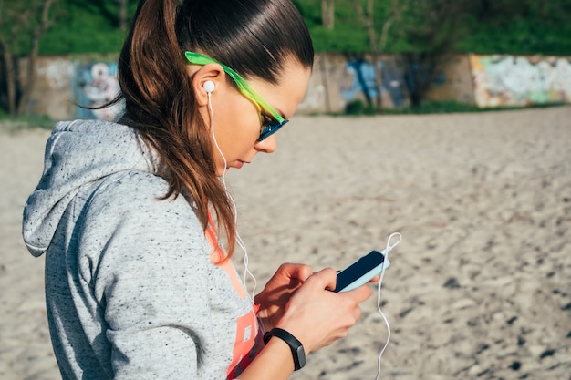 Jeune femme en vêtements de sport avec capuche marchant sur la plage et écoutant de la musique avec des écouteurs sur un téléphone intelligent