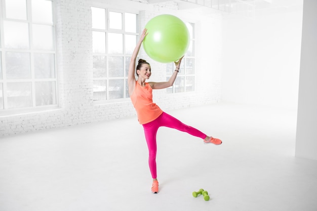 Jeune femme en vêtements de sport colorés qui s'étend avec ballon de fitness dans la salle de sport blanche