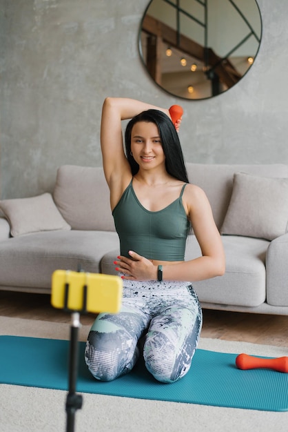 Une jeune femme en vêtements de sport est assise sur le sol avec des haltères et utilise un smartphone sur un trépied à la maison dans le salon pour des leçons vidéo Le concept de sports et de loisirs