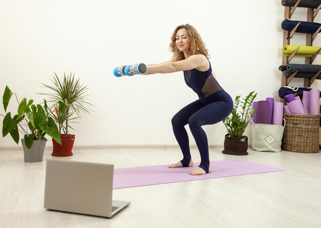 Jeune femme en vêtements de sport faisant des exercices de squat avec des haltères et regardant des vidéos sur un ordinateur portable pendant l'entraînement physique