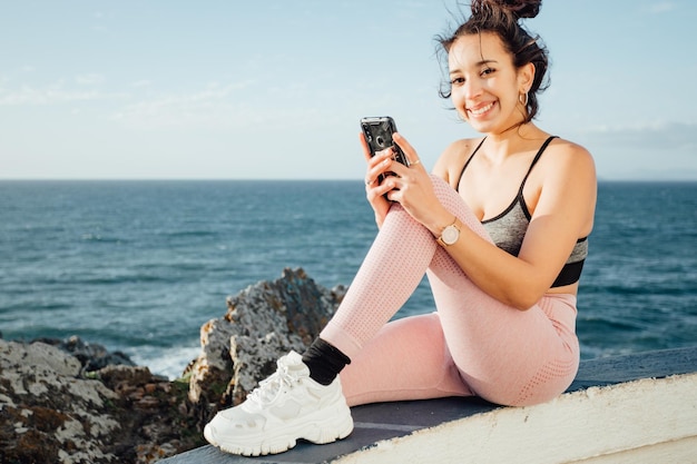 Jeune femme en vêtements de sport vérifiant son smartphone à l'extérieur après une journée de trail sur le rivage d'un lieu naturel Sur le rivage pendant une journée ensoleillée, venteuse et ambiante naturelle