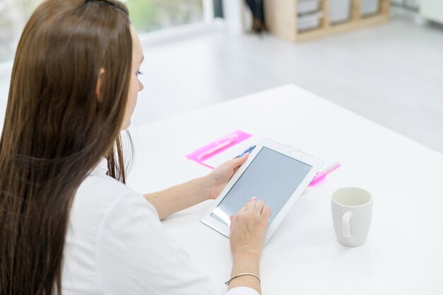 Une jeune femme vêtue d'une chemise blanche et d'un jean est assise à une table avec une tablette dans les mains Une femme chef de bureau travaille sur un appareil moderne La vue de derrière