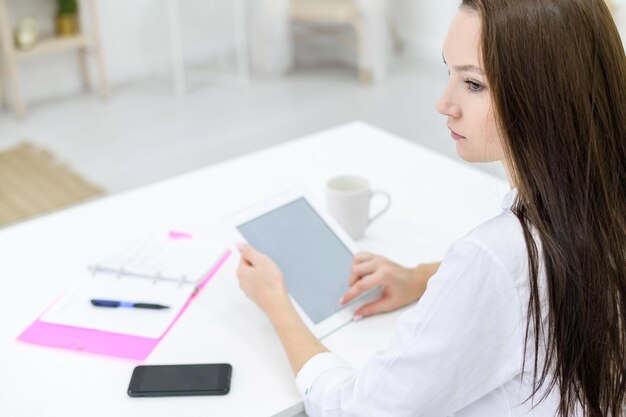 Une jeune femme vêtue d'une chemise blanche et d'un jean est assise à une table avec une tablette dans les mains Une femme chef de bureau travaille sur un appareil moderne La vue de derrière