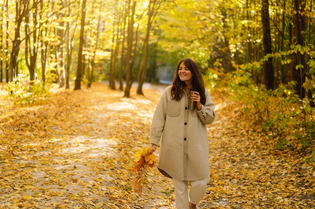 Une jeune femme vêtue d'une longue chemise avec une tasse thermo dans ses mains se promène dans la forêt d'automne