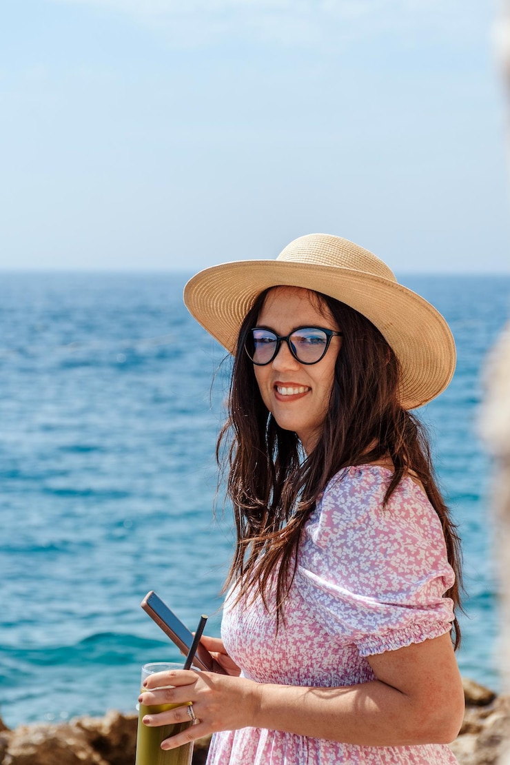Photo une jeune femme vêtue d'un maillot de soleil se tient au bord de la mer en utilisant un téléphone portable et une boisson en main