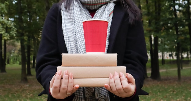 Une jeune femme vêtue d'un manteau noir et d'une écharpe grise tient une pile de livres et un verre avec du café dans les mains sur le fond d'un parc en automne. En plein air. Éducation, fermer les mains de la fille.