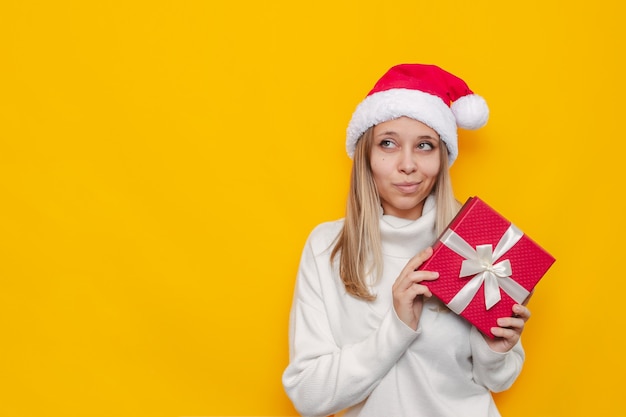 Une jeune femme vêtue d'un pull blanc et d'un chapeau de père Noël en détournant les yeux vers l'espace de copie avec un cadeau