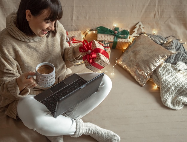 Une jeune femme vêtue d'un pull douillet achète des cadeaux de Noël sur Internet. Concept de choix de cadeaux en ligne et à distance.