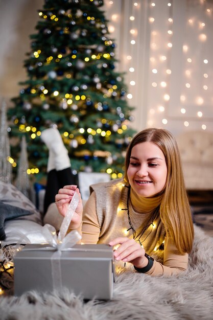 Une jeune femme vêtue d'un pull avec de longs cheveux magnifiques s'assoit et rit devant des cadeaux de Noël. Bonne année